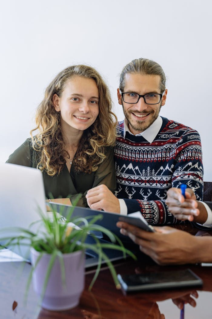about-us Smiling couple with curly hair and eyeglasses using technology at worktable with plant.